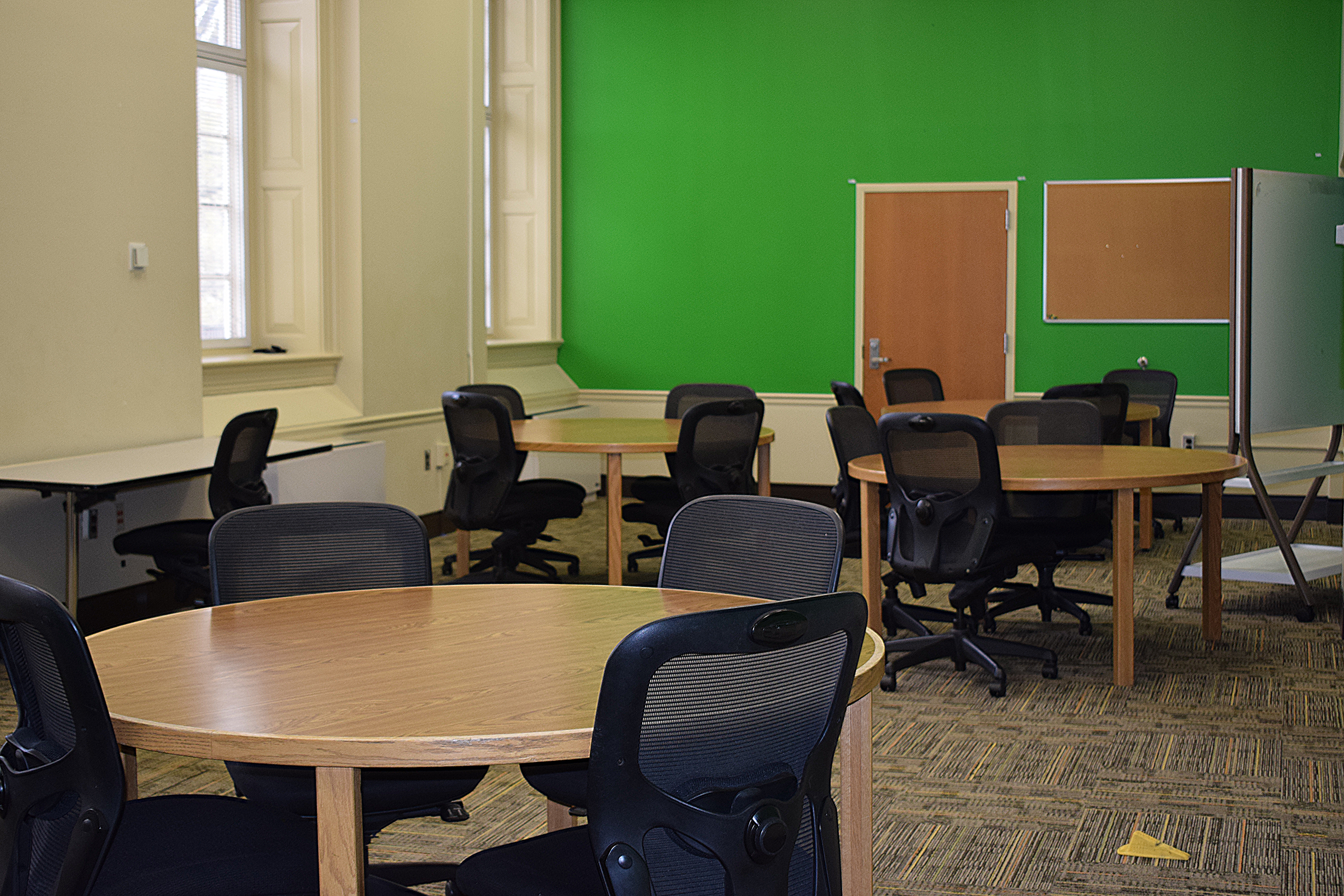 A room with three circular tables surrounded by chairs, a rolling whiteboard, and long, tall windows