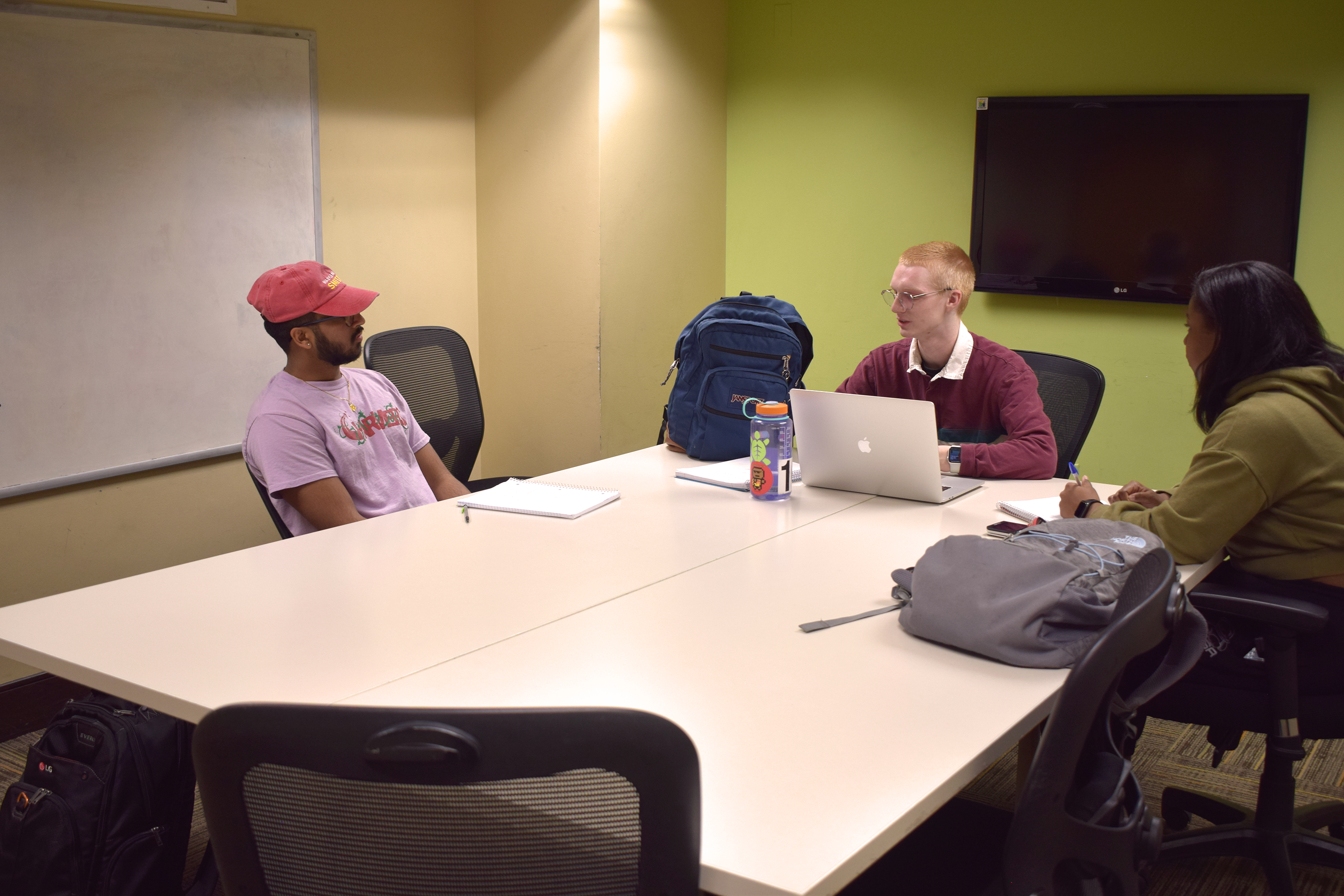 Three students sitting around a table having a discussion. A large monitor and a whiteboard are mounted on the walls.