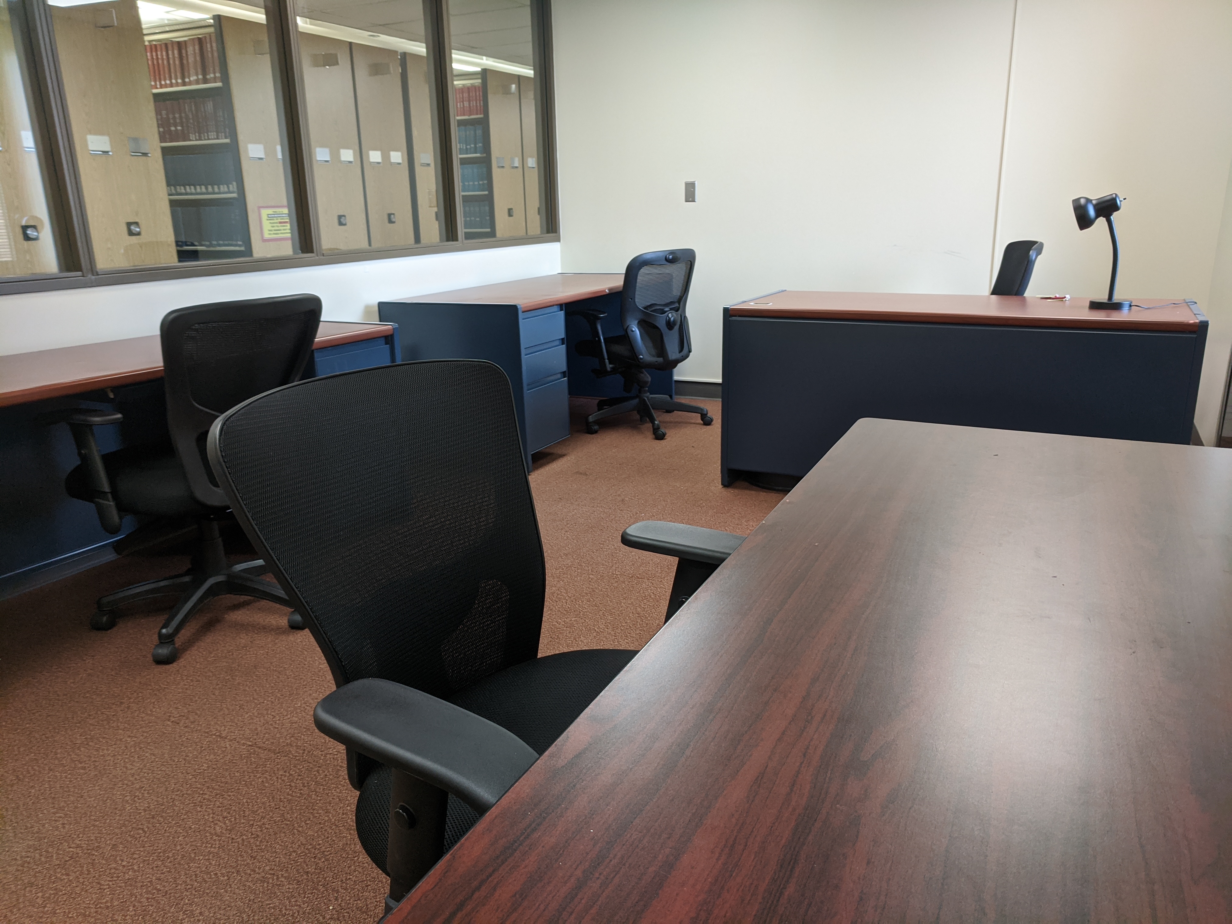 Three empty desks, each with desk chairs, as well as an empty table with one chair visible. One desk has a lamp on it, and one wall of the room is partially a window looking into the library with a view of bookshelves