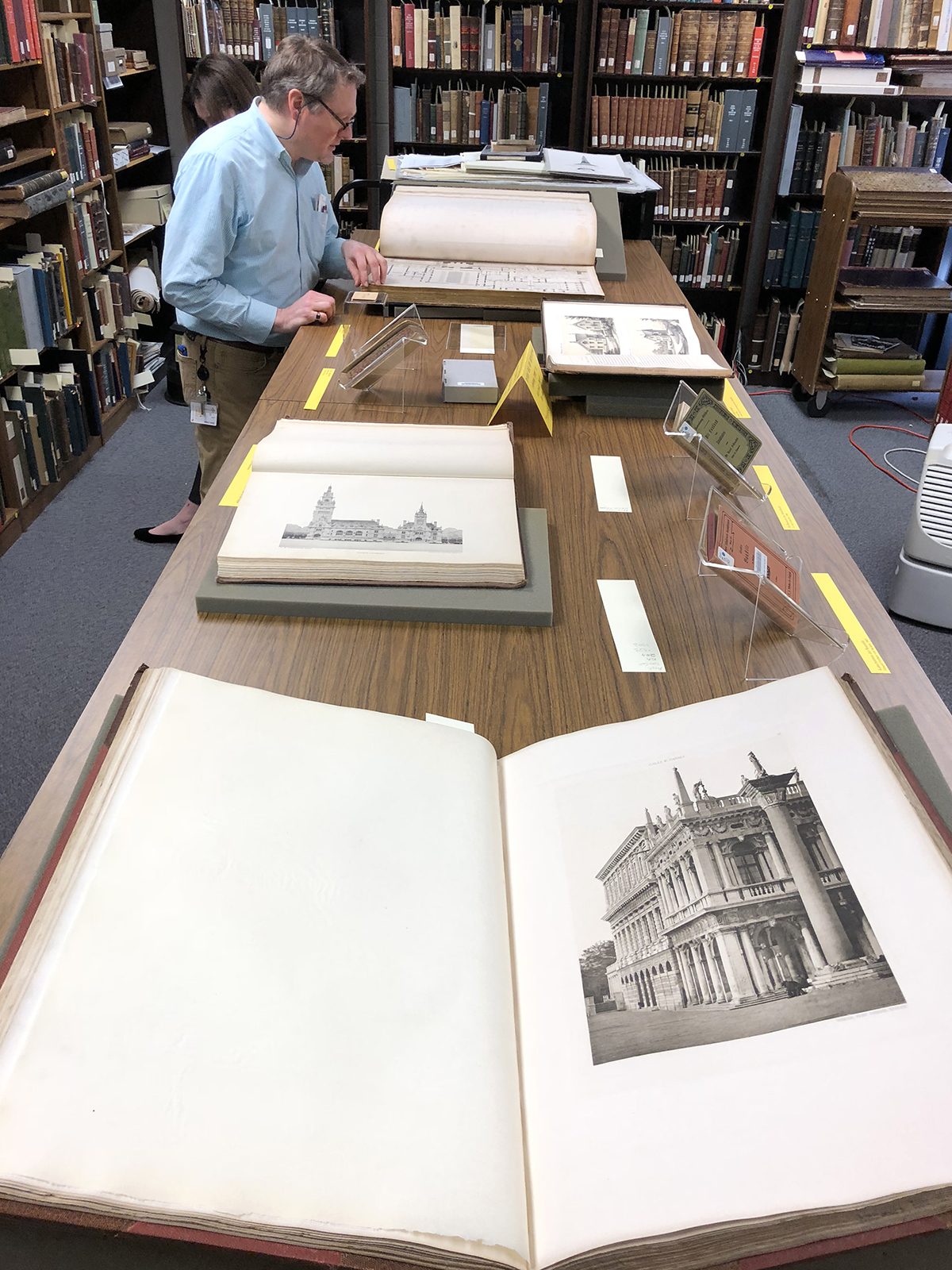 Architecture book open on a table top, with book shelves in the background. Person standing at the table.