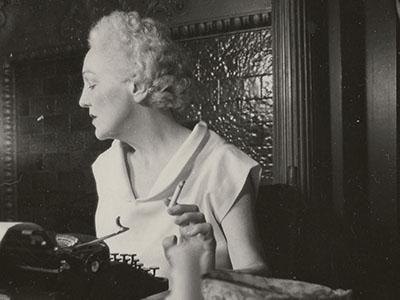 Katherine Anne Porter holding a cigarette in her apartment, circa July 1950-May 1951
