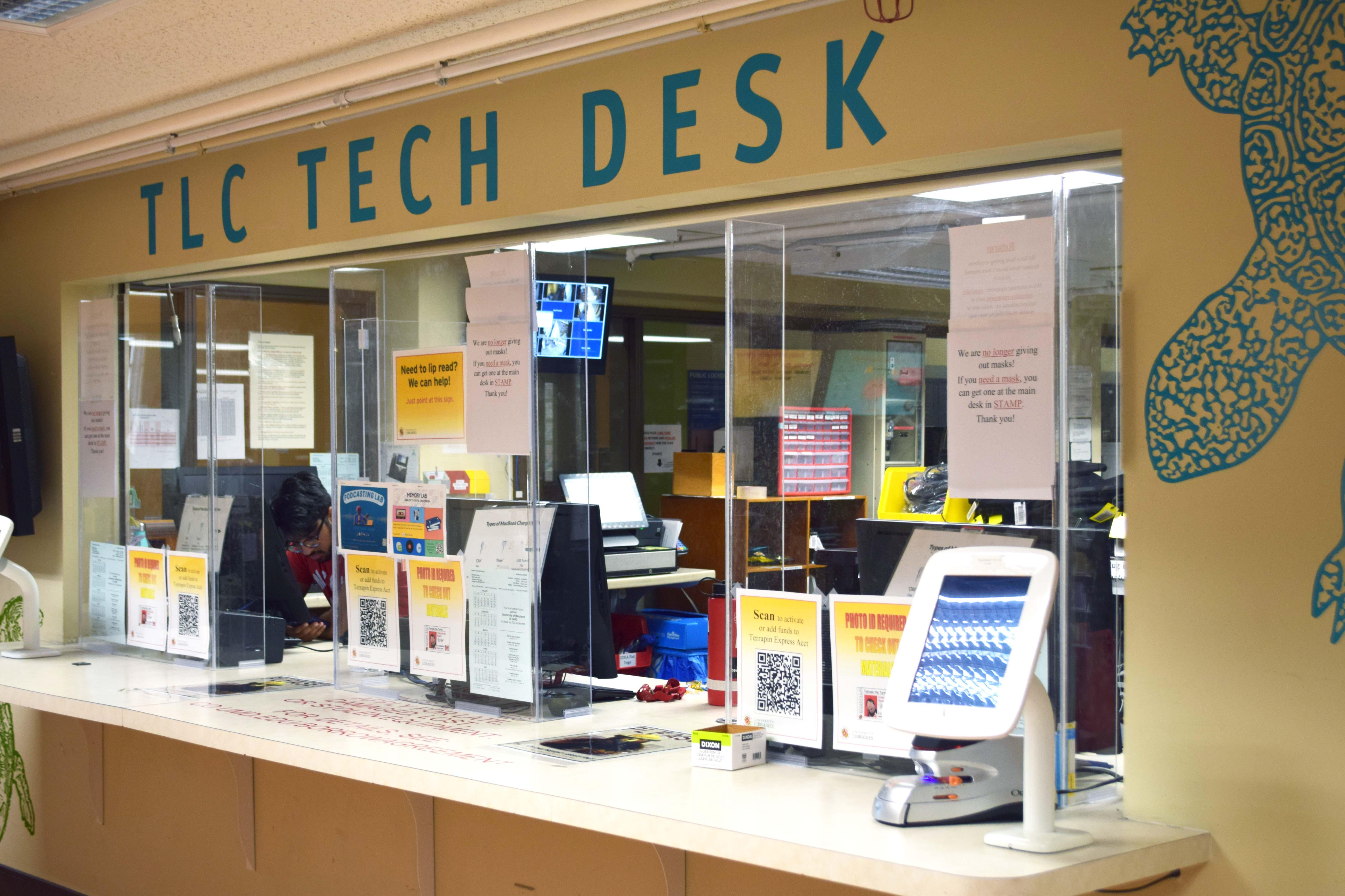 Photo of the TLC Tech Desk from the front. There are computers set along the desk with student workers sitting at them. The open space between the workers and where people approach the desk has plexiglass shielding with some gaps in it. 