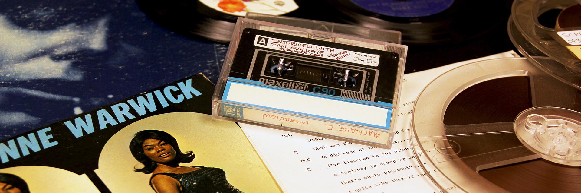Records by Dionne Warwick and Joni Mitchell laid on a table with a 1/4" tape and a cassette of an interview with Ian MacKaye