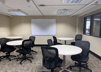 Four small circular tables, each with a few chairs around it. The chairs are wheeled and armless with mesh backs. There is a whiteboard on the wall and a window looks out into the interior of the library.