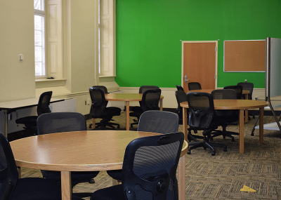 Three circular tables surrounded by wheeled armless chairs with mesh backs in a spacious room with tall windows and a green wall.