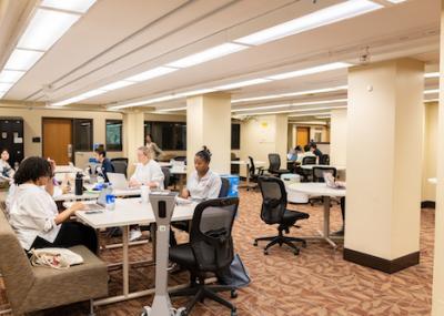 An open area of the library with tables of different sizes and shapes spread around, accompanied by seating ranging from wheeled office chairs to padded benches. Many students seated at the tables are working on laptops, and a charging stand on wheels is visible near one of the tables.