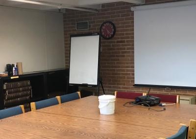 Room with a large square table, padded chairs, white board and projector screen