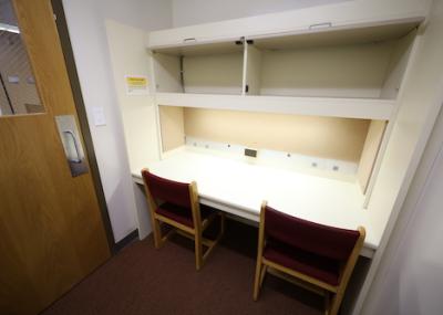 A small room with one wall dominated by a desk topped with shelving. Two wooden padded chairs are placed at the desk and the closed door holds a small window looking out into the library.