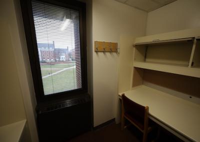 Small room with a desk and shelves along one wall. There is a wooden padded chair at the desk and a coat rack mounted on the wall next to a window with blinds.