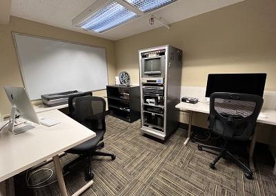 Three rectangular work tables with Apple computers and mesh backed office chairs. There is a white board on one wall and audio visual media converting equipment against another