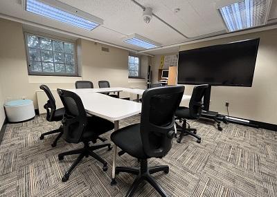 Carpeted room with a U-shape table surrounded by office chairs, facing a large screen. There  is a computer in one corner and two windows on one wall.