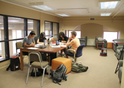 Carpeted room with large windows along one wall. There is a group of students sitting working at a large table and armchairs around the room.