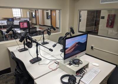 Three rectangular tables arranged together with an Apple Computer and headphones and microphones. There are two windows that look out onto the communal computer area.