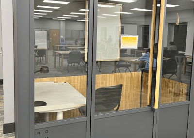 tall rectangular booth with three windows paneling the front. The middle window has a long door handle running vertically down it's right side. Inside on the left there is desk with a desk lamp, a padded office chair and a small whiteboard mounted on the back wall.