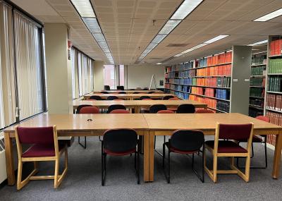 Carpeted windowed space next to book shelves, over head lit with long wooden tables and padded chairs configured in rows with a whiteboard easel.