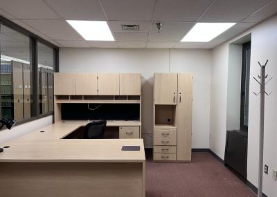 A large wooden desk with storage shelves and cabinets facing a wide window into the library. The desk has a small lamp on it and nearby is a compact storage unit with drawers and a narrow closet.