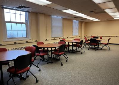 Large carpeted room with windows and outlets along one wall, there is a row of round tables with cushioned desk chairs grouped around them. The chairs have wheels on the bottom and a cushioned back.