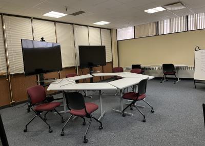 Carpeted room with small tables arranged into a larger hexagon face two standing monitors. There are backed cushioned chairs with wheels grouped around the table formation. Along the back wall are two individual tables with chairs and a whiteboard easel.