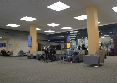 Students sitting in low padded chairs in a carpeted open area with overhead lighting. There are low tables staggered throughout the space