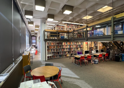  Carpeted open area with a tall ceiling a row of individual tables and chairs facing the window wall. In the foreground there is a printer and a round table with four chairs group around it. Also in view are four Mac computers on tables with chairs in a row, and a book shelf behind. A staircase to the right of the computers is visible leading up to the second floor that wraps around the space and includes more individual study tables and book shelves. 