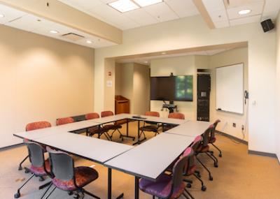 Carpeted room with six tables and twelve chairs arranged in a rectangle, a monitor, window, and large whiteboard